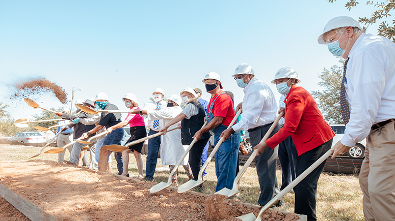 A group of thirteen men and women in a line with shovels and wearing hardhats at a ground breaking for a new construction project